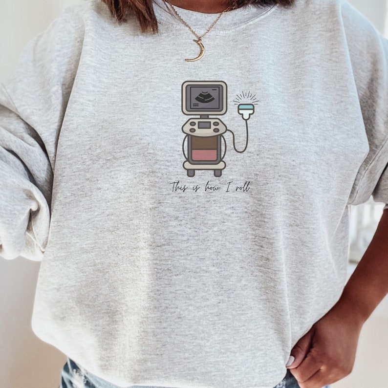 May include: A light gray sweatshirt featuring a cartoon ultrasound machine graphic. The machine is brown, beige, and pink, with a screen displaying an image. The text "This is how I roll" is printed below the graphic.