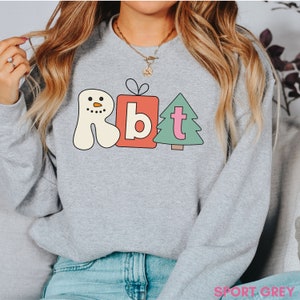 May include: A light gray sweatshirt with a colorful graphic featuring a snowman, a gift, and a Christmas tree. The graphic has the letters "Rbt" in a playful font. The sweatshirt is made of a soft material.
