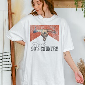 May include: White t-shirt with a distressed red graphic of a bull skull and the text "Raised On 90's Country".
