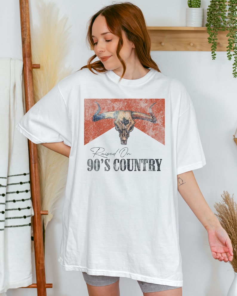May include: White t-shirt with a distressed red graphic of a bull skull and the text "Raised On 90's Country".