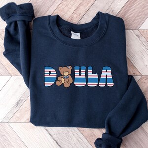 May include: Navy blue sweatshirt with the word "DOULA" in white letters with red and blue stripes. A brown teddy bear with a blue bow is in the middle of the word.