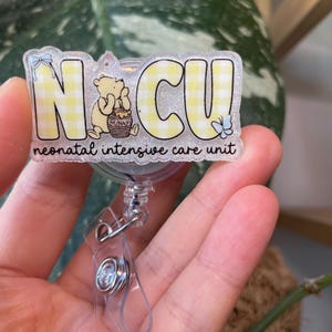 May include: A badge reel with the letters "NCU" in yellow gingham print, featuring a cartoon bear and the words "neonatal intensive care unit". The reel has a clear plastic clip and a retractable cord.