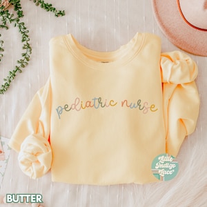 Puede incluir: Una sudadera amarilla con el texto "pediatric nurse" escrito en letras de colores. La sudadera está doblada y colocada sobre una superficie blanca.