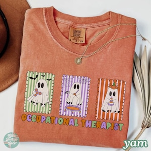 May include: Orange Comfort Colors t-shirt with a graphic of three cartoon ghosts in frames. The frames have striped backgrounds in green, purple, and orange. The text "OCCUPATIONAL THERAPIST" is below the frames.
