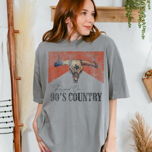 May include: A gray t-shirt with a vintage-style graphic of a bull skull with a red, white, and blue bandana. The text "Raised On 90's Country" is printed below the graphic.