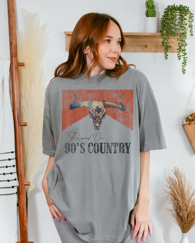 May include: A gray t-shirt with a vintage-style graphic of a bull skull with a red, white, and blue bandana. The text "Raised On 90's Country" is printed below the graphic.