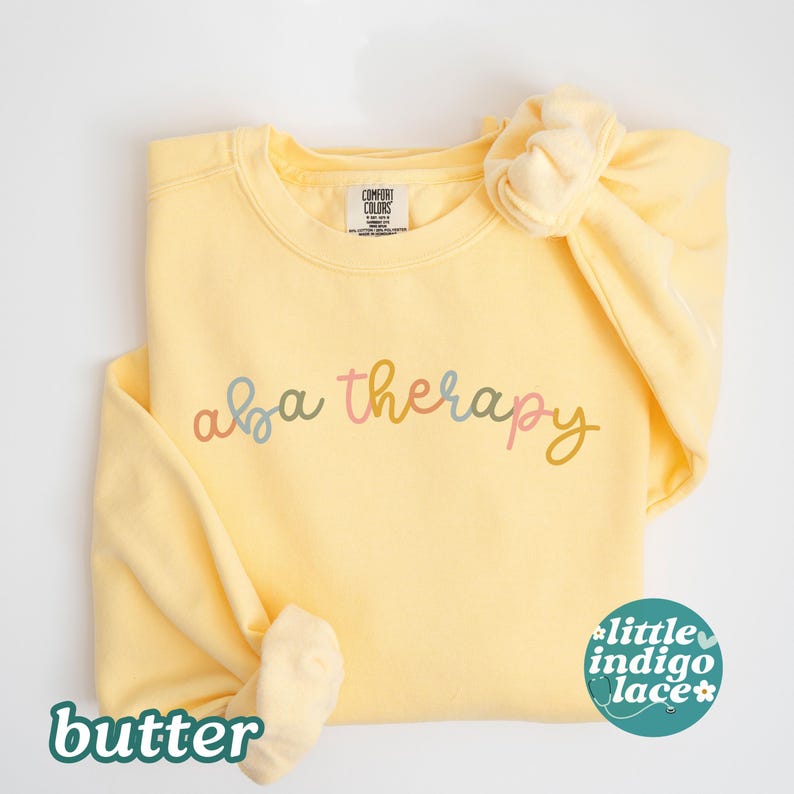 K&ouml;nnte beinhalten: Ein hellgelbes Sweatshirt mit den Worten "aba therapy" in Pastellfarben. Ein passendes Scrunchie befindet sich auf der Schulter. Das Wort "butter" befindet sich unten links und ein kleines Logo unten rechts.