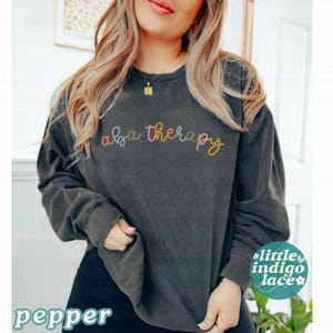 K&ouml;nnte beinhalten: Dunkelgraues Sweatshirt mit dem Schriftzug "aba therapy" in farbenfroher Schrift auf der Brust. Das Sweatshirt hat lange &Auml;rmel und einen Rundhalsausschnitt. Das Wort "pepper" ist unten links aufgedruckt.