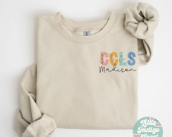 Op de afbeelding: Een zandkleurig sweatshirt met de tekst "CCLS Madison" in kleurrijke letters met bloemmotieven. Het sweatshirt is gevouwen en gefotografeerd op een witte achtergrond.