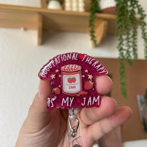 Puede incluir: Portagafete de purpurina rosa con un dise&ntilde;o de tarro de mermelada de fresa y el texto "Occupational Therapy is my jam".