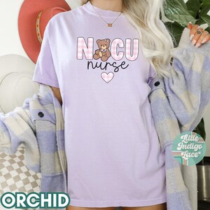 Puede incluir: Una camiseta de color morado claro con un dise&ntilde;o a cuadros rosa y blanco. La camiseta presenta un osito de peluche de dibujos animados y el texto "NCU nurse" con un coraz&oacute;n rosa.