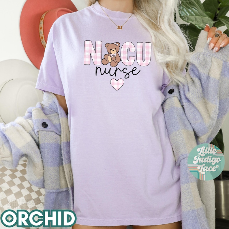 Puede incluir: Una camiseta de color morado claro con un dise&ntilde;o a cuadros rosa y blanco. La camiseta presenta un osito de peluche de dibujos animados y el texto "NCU nurse" con un coraz&oacute;n rosa.