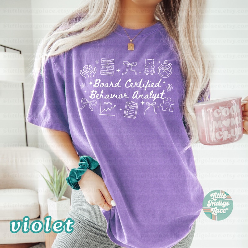May include: A violet-colored t-shirt with the text "Board Certified Behavior Analyst" in white. The shirt features various small graphic icons. A pink mug with the text "Cool Mom Club" is held in the hand.