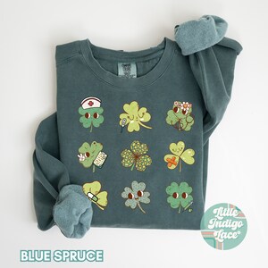 K&ouml;nnte beinhalten: Ein blaues Sweatshirt mit einem Motiv aus neun vierbl&auml;ttrigen Kleebl&auml;ttern, jedes mit einem anderen medizinischen Design. Die Kleebl&auml;tter haben Gesichter und tragen Gegenst&auml;nde wie eine Krankenschwesterm&uuml;tze, einen Verband und ein Thermometer. Der Text "Little Indigo Lace" befindet sich in einem Kreis unten rechts auf dem Sweatshirt.