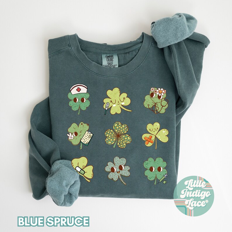 K&ouml;nnte beinhalten: Ein blaues Sweatshirt mit einem Motiv aus neun vierbl&auml;ttrigen Kleebl&auml;ttern, jedes mit einem anderen medizinischen Design. Die Kleebl&auml;tter haben Gesichter und tragen Gegenst&auml;nde wie eine Krankenschwesterm&uuml;tze, einen Verband und ein Thermometer. Der Text "Little Indigo Lace" befindet sich in einem Kreis unten rechts auf dem Sweatshirt.