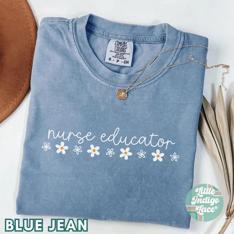 以下が含まれることがあります： 白い文字で「nurse educator」と書いてある、青いジーンズ地のTシャツ。白いデイジーが1列に並んでいます。このTシャツは、使い古したような風合いになっており、Comfort Colors製です。