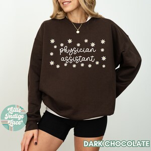 May include: A dark chocolate brown sweatshirt with the text "physician assistant" in white lettering surrounded by white daisy flowers.