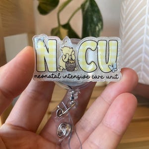 May include: A badge reel with the letters "NCU" in yellow, with a Winnie the Pooh graphic. The text "neonatal intensive care unit" is below the letters. The badge reel is clear with a metal clip.