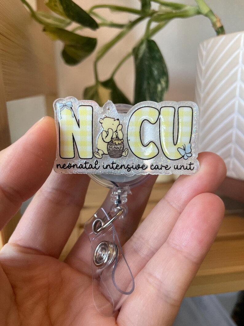 May include: A badge reel with the letters "NCU" in yellow, with a Winnie the Pooh graphic. The text "neonatal intensive care unit" is below the letters. The badge reel is clear with a metal clip.