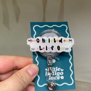 May include: A circular badge reel featuring a white acrylic topper with the black text Child Life, decorated with colorful small shapes like hearts, stars, and flowers. The badge reel is attached to a teal display card with white scalloped borders.