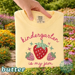May include: A pale yellow t-shirt featuring a graphic of three strawberries with green gingham leaves and small star accents. The text reads "kindergarten is my jam" in a red cursive font. The shirt has a Comfort Colors brand neck label.