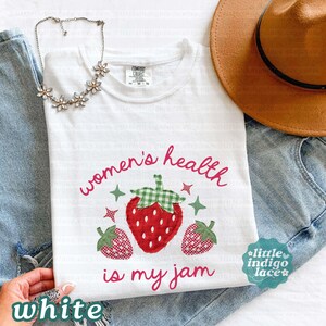 May include: A white t-shirt featuring a graphic of three strawberries with green gingham leaves and the text "women's health is my jam" in pink script. The shirt is paired with blue denim jeans, a brown wide-brimmed hat, and a silver flower necklace.