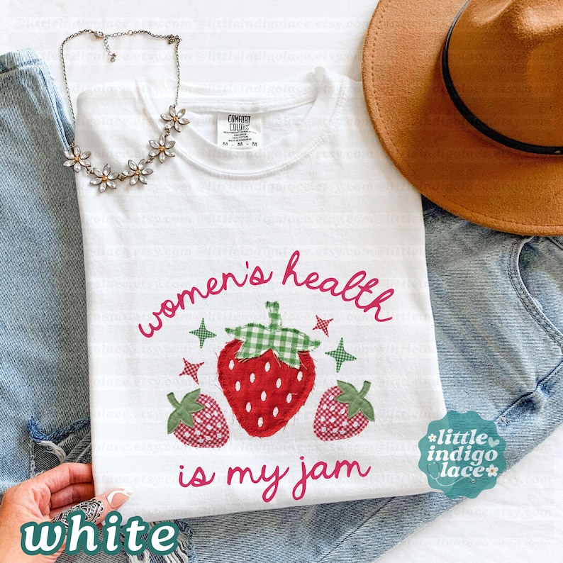 May include: A white t-shirt featuring a graphic of three strawberries with green gingham leaves and the text "women's health is my jam" in pink script. The shirt is paired with blue denim jeans, a brown wide-brimmed hat, and a silver flower necklace.