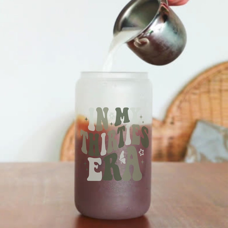 May include: A frosted glass tumbler with the text "IN MY THIRTIES ERA" in a retro font. The tumbler is filled with a dark brown liquid and a silver pitcher is pouring a white liquid into the tumbler.