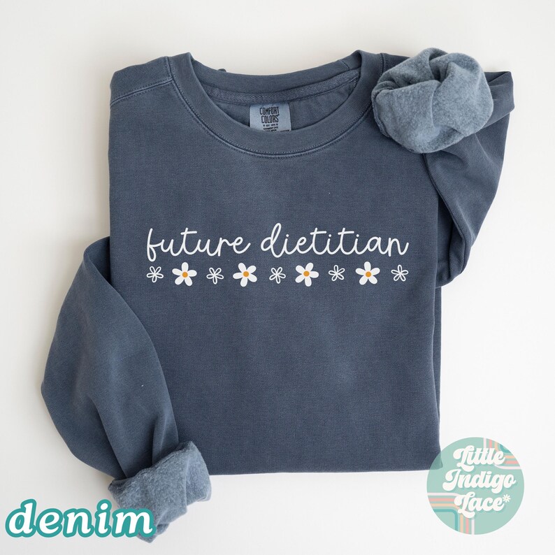 May include: Denim blue sweatshirt with the text "future dietitian" in white script, decorated with white daisy graphics. The sweatshirt has a crew neck and long sleeves. The word "denim" is printed in a teal color at the bottom left.