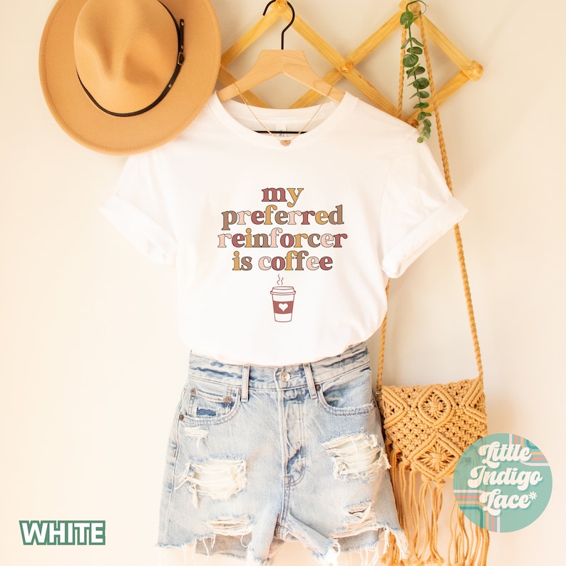 May include: White t-shirt with the text "my preferred reinforcer is coffee" in brown, pink, and beige lettering. A cartoon coffee cup with steam and a heart is below the text.