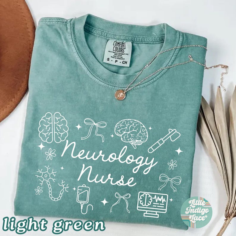 May include: Light green t-shirt with the words "Neurology Nurse" and related medical graphics in white. The shirt has a crew neck and short sleeves. A silver necklace with a pendant is draped across the shirt.