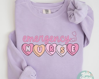 Puede incluir: Una sudadera de color lavanda con el texto "emergency nurse" en letras rosa y blancas. La palabra "nurse" está escrita con cinco corazones de caramelo rosa, cada uno con una letra diferente. Un estetoscopio está dibujado junto a la palabra "emergency".