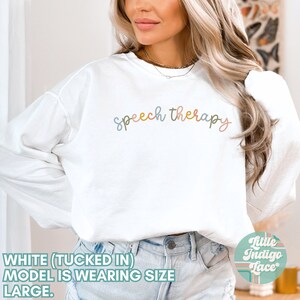 May include: White long-sleeved sweatshirt with the text "speech therapy" in colorful script. The model is wearing a size large and has the shirt tucked in.