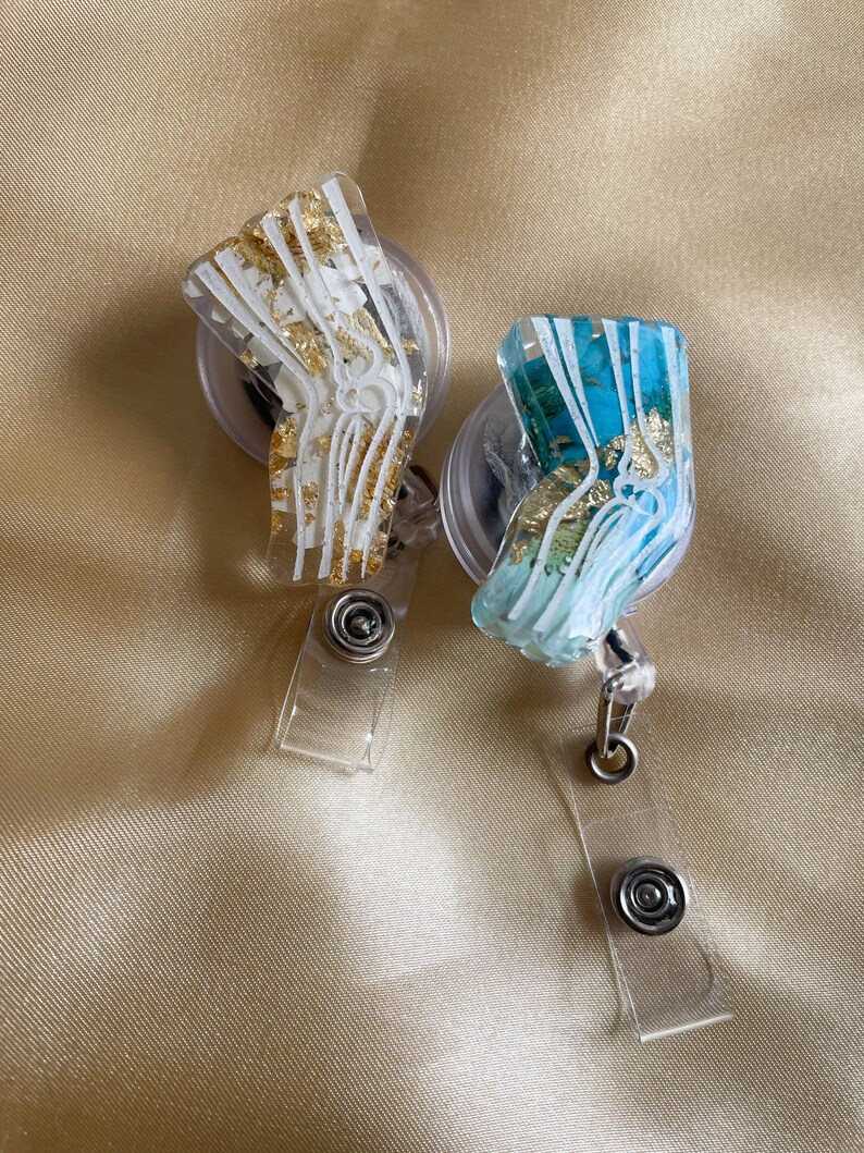 Pu&ograve; includere: Due porta badge in plastica trasparente con un design di osso anatomico in oro e bianco. Un porta badge ha uno sfondo bianco con brillantini dorati e l'altro ha uno sfondo blu con brillantini dorati.