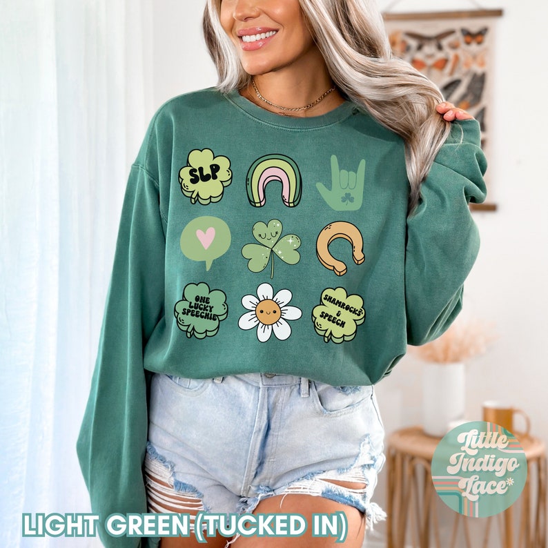 May include: A light green sweatshirt with a variety of St. Patrick's Day themed graphics, including shamrocks, rainbows, a horseshoe, and the text "SLP" and "One Lucky Speechie". The sweatshirt is tucked in and the model is wearing denim shorts.