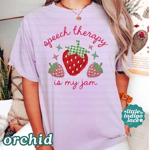 K&ouml;nnte beinhalten: Lilafarbenes T-Shirt mit dem Schriftzug "speech therapy is my jam" in rosa Schrift. Das Shirt zeigt eine gro&szlig;e Erdbeer-Grafik mit einem gr&uuml;n karierten Oberteil und zwei kleineren Erdbeer-Grafiken.