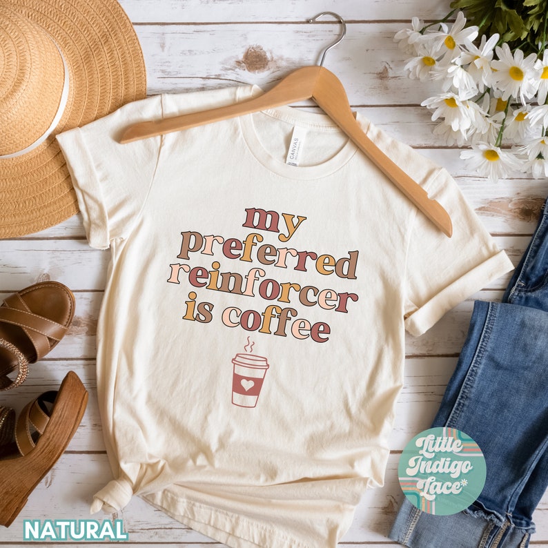 May include: A beige t-shirt with the text "my preferred reinforcer is coffee" in brown, pink, and orange lettering. A cartoon coffee cup with a heart is below the text. The shirt is on a hanger and has a "NATURAL" label at the bottom.