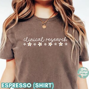 May include: A brown t-shirt with the text "clinical research" in white lettering and a row of white daisies below the text. The shirt is being worn by a person with long blonde hair. The text "ESPRESSO (SHIRT)" is at the bottom of the image.