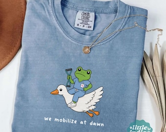 May include: A blue jean colored t-shirt featuring a graphic of a green frog wearing blue scrubs and holding a medical tool while riding on the back of a white goose. The text below the graphic reads, we mobilize at dawn.
