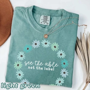 May include: Light green Comfort Colors t-shirt with a daisy chain design and the text "see the able not the label." A silver necklace with a pendant rests on the shirt. The shirt is labeled "light green."