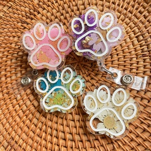 May include: Four clear paw print shaped keychains with white outlines. Each keychain has a different colour background and is filled with dried flowers and gold flakes.