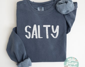 May include: A dark blue crewneck sweatshirt featuring the word SALTY printed in white distressed lettering across the chest. The garment has a visible interior fleece lining and a Comfort Colors brand label at the inner neckline.
