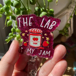 May include: A glittery dark pink badge reel featuring a central illustration of a strawberry jam jar with a red and white checkered lid. The text reads THE LAB IS MY JAM in white lettering, surrounded by small yellow stars and strawberry icons.