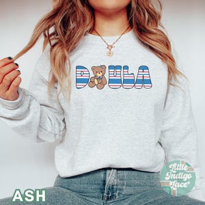 May include: A gray sweatshirt with the word "DOULA" in blue and red striped letters. A small teddy bear with a blue bow tie is in the middle of the word. The sweatshirt is being worn by a person with long brown hair.