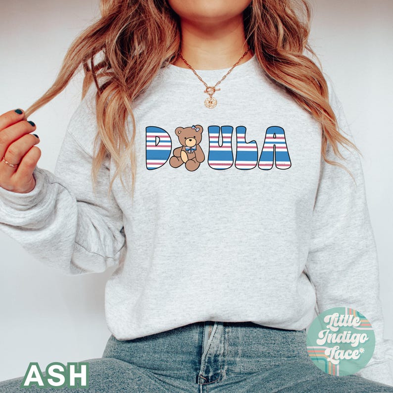 May include: A gray sweatshirt with the word "DOULA" in blue and red striped letters. A small teddy bear with a blue bow tie is in the middle of the word. The sweatshirt is being worn by a person with long brown hair.