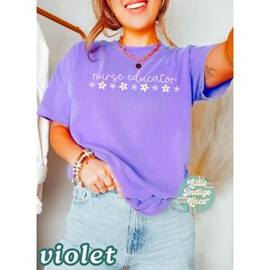 以下が含まれることがあります： 紫色のTシャツに「nurse educator」という文字と、白い花が1列にプリントされています。Tシャツは、金色のネックレスとブレスレットをしている人が着用しています。画像の下部に緑色の「violet」という文字がプリントされています。