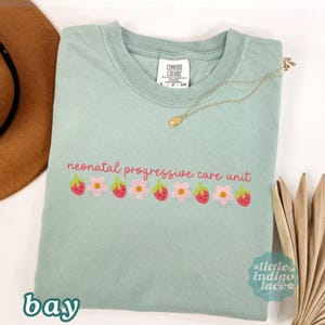 May include: A light green t-shirt with the words "neonatal progressive care unit" in pink script, decorated with strawberry and flower graphics. The shirt has a crew neck and short sleeves. A gold necklace is draped across the shirt.