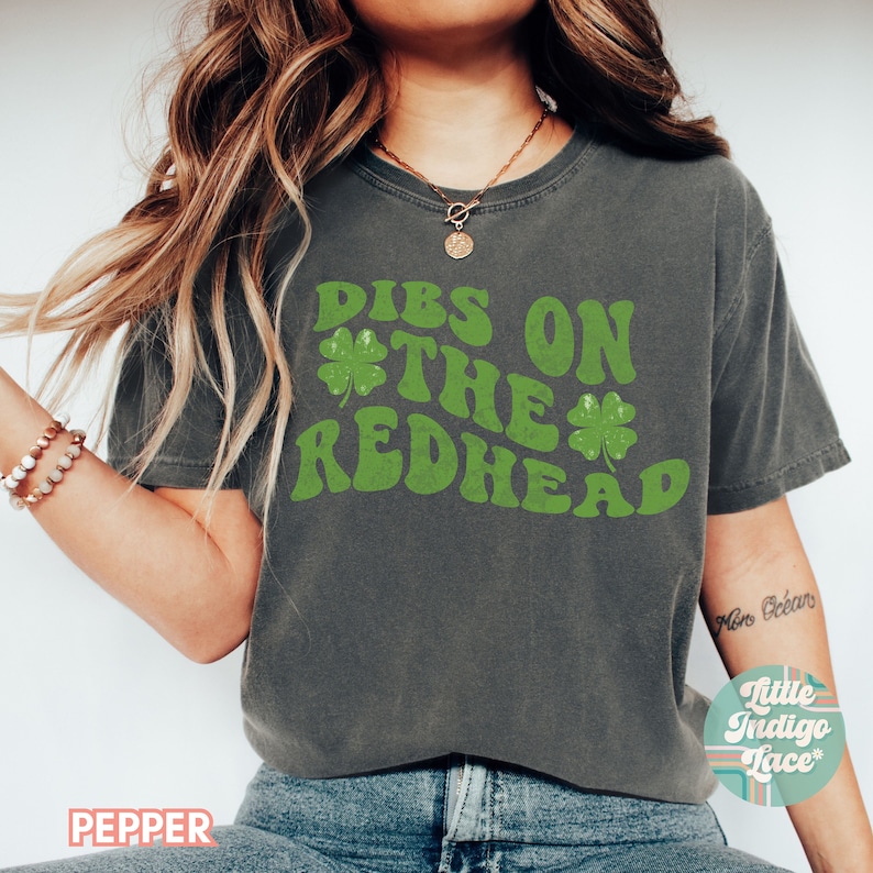 Op de afbeelding: Een donkergrijs T-shirt met een groene afbeelding waarop "DIBS ON THE REDHEAD" staat met klavertjesvier designs.