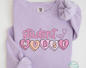 May include: A lavender sweatshirt with the text "Student Nurse" in pink letters. The letters are surrounded by pink and white candy hearts. A stethoscope is drawn above the text.