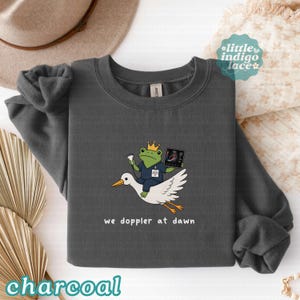May include: A charcoal gray crewneck sweatshirt featuring a graphic of a green frog wearing a gold crown and blue scrubs, holding an ultrasound probe and monitor while riding a white stork, with the text we doppler at dawn printed below.
